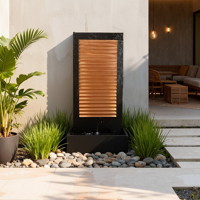 Modern rectangular wall fountain with copper horizontal slats and dark base, positioned beside patio walkway and greenery.