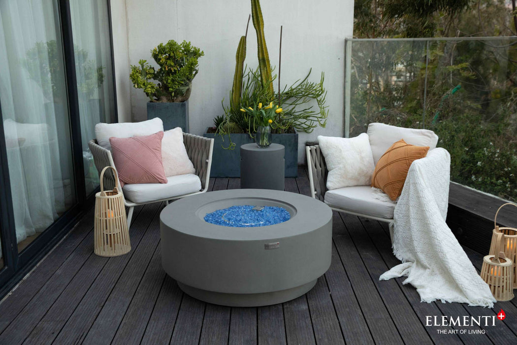 A modern round concrete Colosseo fire pit table on a deck, featuring white furniture, ideal for enhancing outdoor relaxation