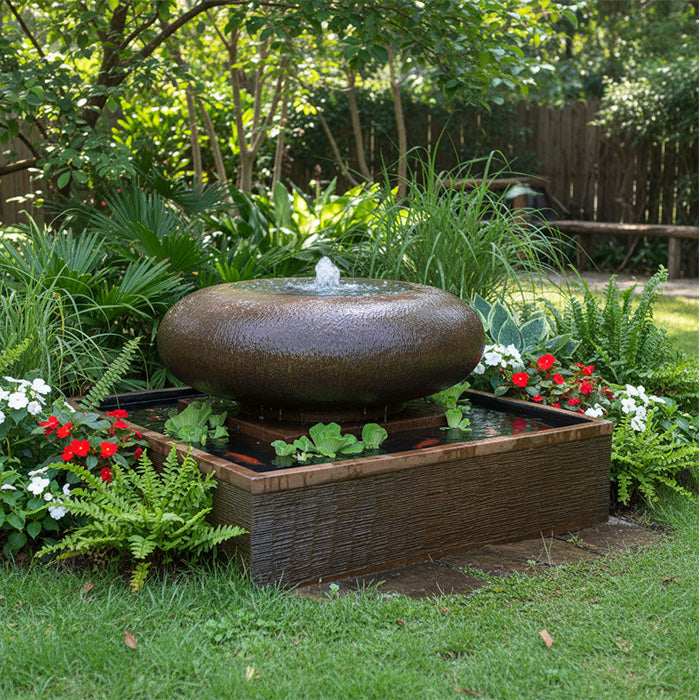 Contemporary moon bowl garden water fountain surrounded by colorful flowers and greenery in landscaped backyard setting.