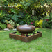 Round moon bowl outdoor water fountain in square basin with floating plants, set in lush green garden landscape with tropical foliage.