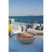 An unlit moon gray concrete fire bowl filled with reddish-brown lava rock sits on a painted blue wood deck overlooking the open ocean, flanked by white outdoor lounge chairs and a mustard yellow throw blanket in bright daylight.