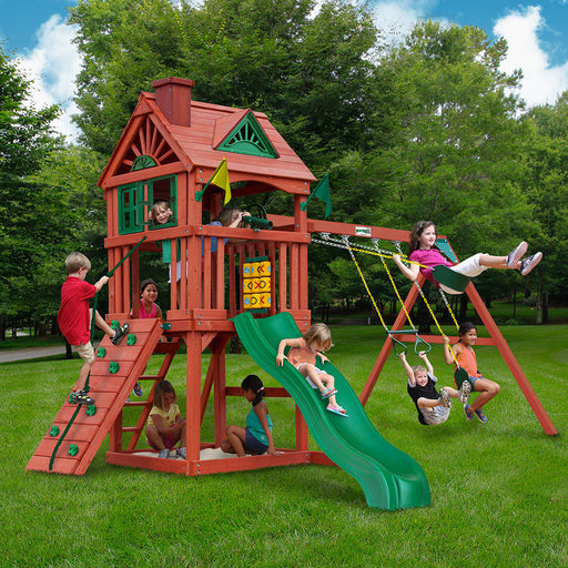 Kids playing on the Gorilla Playsets Nantucket II with treehouse roof, green slide, rock wall, sandbox, and swing set in a backyard