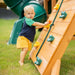 Toddler boy climbing the wooden rock wall with colorful hand grips and nylon rope on a Gorilla Playsets swing set in a grassy backyard
