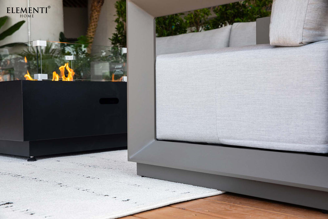 Close-up of the Elementi Outdoor Furnitures Newport Collection with a fire pit table with a glass wind guard, on a white rug.