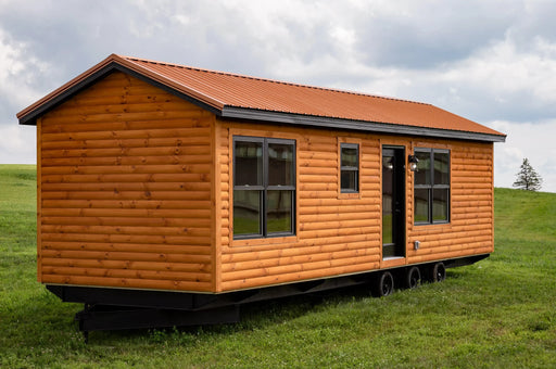 Wildfire Cabin with log siding and a metal roof. It has several windows and a black door. It's situated on a trailer in a green field with a cloudy sky.