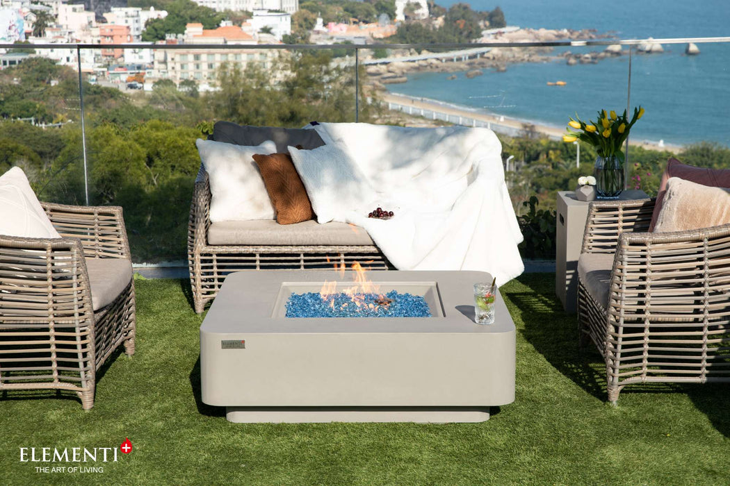 Outdoor furniture arranged on a patio with the Elementi Plus Lucerne Square Concrete Fire Pit Table, showcasing an ocean vista.