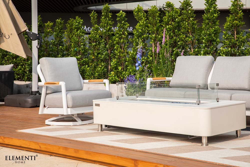 A light gray Elementi Santorini Collection outdoor armchair and a white rectangular fire table with a glass wind guard on a wooden patio.