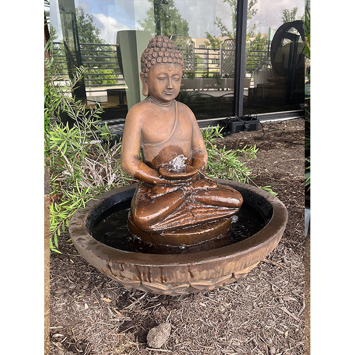Outdoor Exaco Buddha Fountain placed in a landscaped garden area with reflective glass windows in the background, showing peaceful water flow from the statue’s hands.