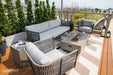 An inviting outdoor living space with Elementi Outdoor Furnitures Milano Collection seating, a fire pit, and a patterned rug on a wooden deck.