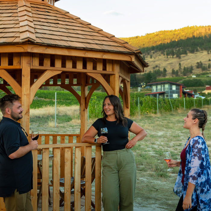 Friends with wine gathered at the Outdoor Living Today 10' Bayside Panelized Octagon Gazebo, cedar shake roof and railings beside a vineyard