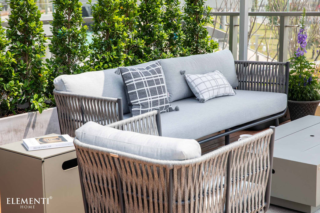 An Elementi Milano Collection outdoor sofa with light grey cushions and two decorative pillows, one with a grey grid pattern and the other a checkered pattern with tassels. 