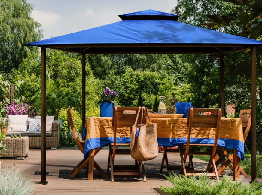 Bright blue canopy of the Kingsbury Soft Top Gazebo providing shade over a cozy wooden deck dining area, surrounded by lush greenery