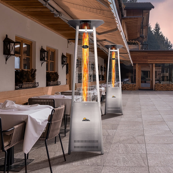 A tall, stainless steel outdoor flame heater by Paragon Outdoor Vesta stands between a dining area with cushions, adding comfort and elegance to the garden scene.