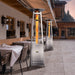 A tall, stainless steel outdoor flame heater by Paragon Outdoor Vesta stands between a dining area with cushions, adding comfort and elegance to the garden scene.