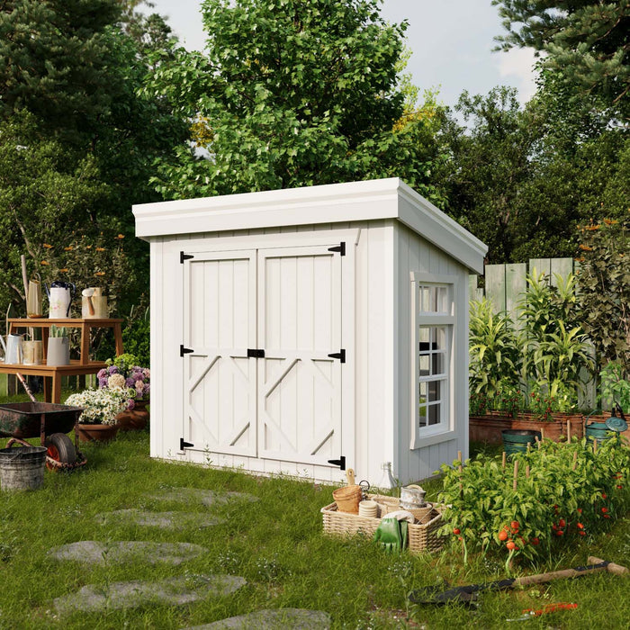 White Petite Greenhouse by Little Cottage Co. placed in a garden setting, surrounded by flowers, vegetables, and gardening tools for backyard use