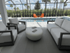Outdoor living set with gray sofa, two lounge chairs and the Elementi Hawaii Pebble cream white table, pool and palm trees beyond