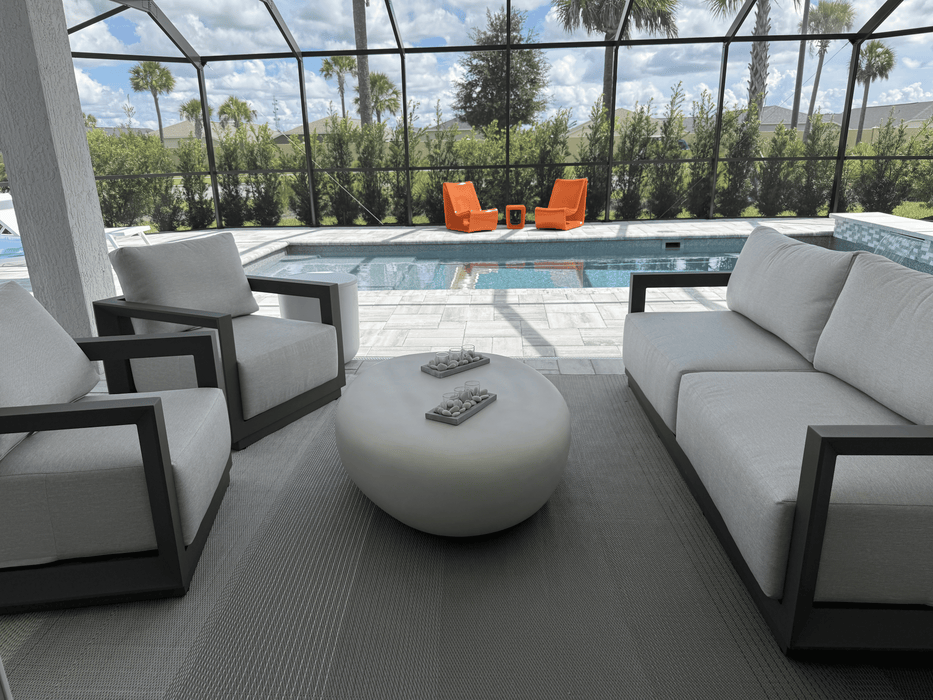 Outdoor living set with gray sofa, two lounge chairs and the Elementi Hawaii Pebble cream white table, pool and palm trees beyond