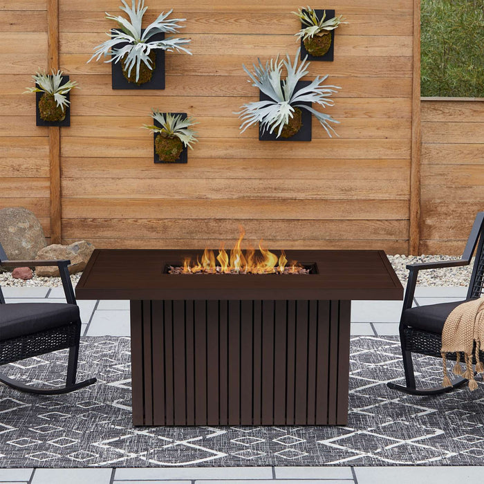 Real Flame Calvin Fire Pit Table bronze burning outdoors on a patio flanked by two black rocking chairs with cushions and a wood slat privacy wall