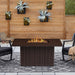 Real Flame Calvin Fire Pit Table bronze burning outdoors on a patio flanked by two black rocking chairs with cushions and a wood slat privacy wall