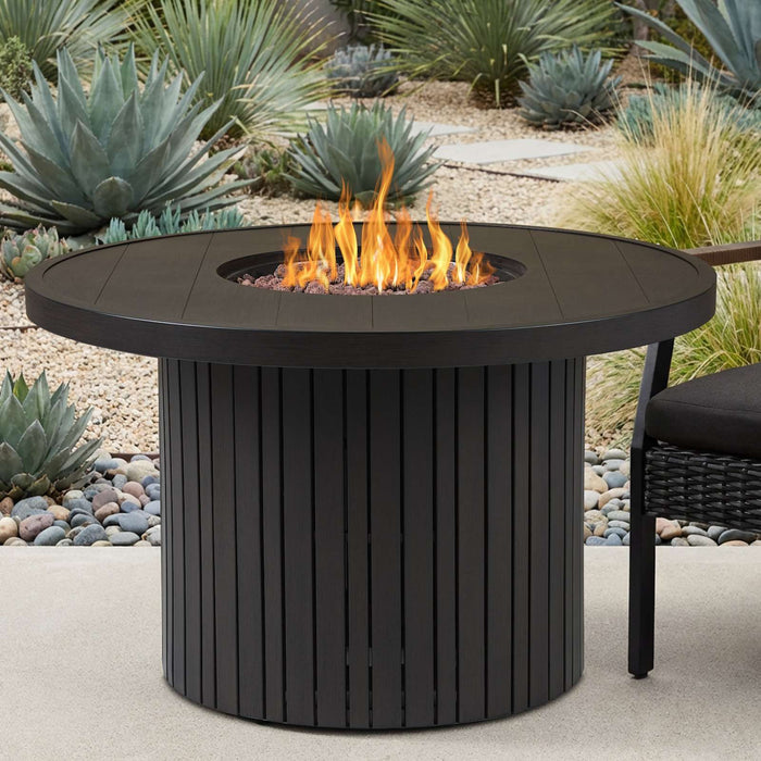 Real Flame Calvin Round Fire Pit Table centered in a landscaped patio with four black cushioned wicker chairs, birch trees, and a modern wood-and-glass home in the background