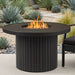 Real Flame Calvin Round Fire Pit Table centered in a landscaped patio with four black cushioned wicker chairs, birch trees, and a modern wood-and-glass home in the background