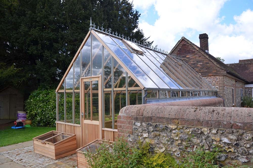 Rear angle of the cedar Westminster greenhouse showing its glass roof, wooden panels, and a brick wall surrounding the garden area.