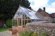 Rear angle of the cedar Westminster greenhouse showing its glass roof, wooden panels, and a brick wall surrounding the garden area.