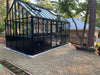 Side garden view of the Retro Royal Victorian VI Greenhouse with black frame and decorative panel design, situated on a gravel foundation among tall pine trees.