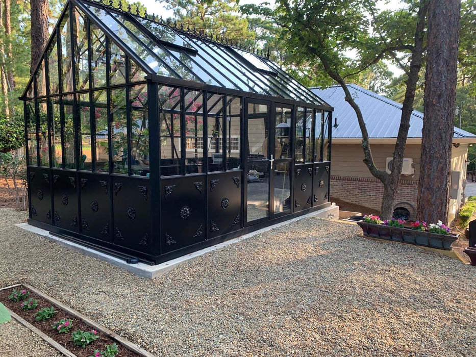 Side garden view of the Retro Royal Victorian VI Greenhouse with black frame and decorative panel design, situated on a gravel foundation among tall pine trees.