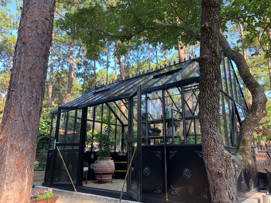 Outdoor side view of the Retro Royal Victorian VI Greenhouse with black frame, decorative lower panels, glass roof, and garden surroundings under tall pine trees.