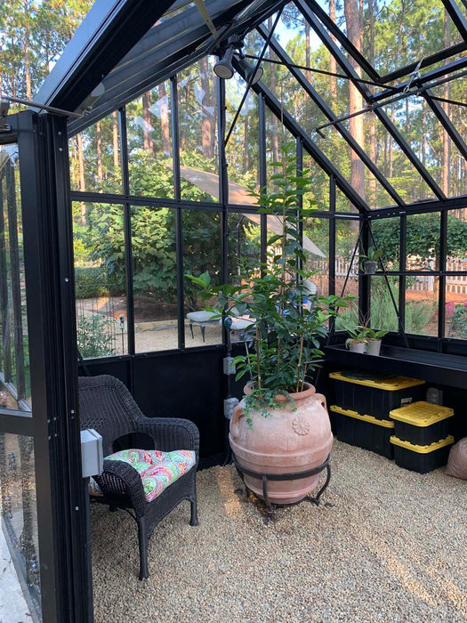 Inside view of the Retro Royal Victorian VI Greenhouse with black frame, terracotta potted plant, storage bins, and scenic garden surroundings visible outside.