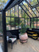 Inside view of the Retro Royal Victorian VI Greenhouse with black frame, terracotta potted plant, storage bins, and scenic garden surroundings visible outside.