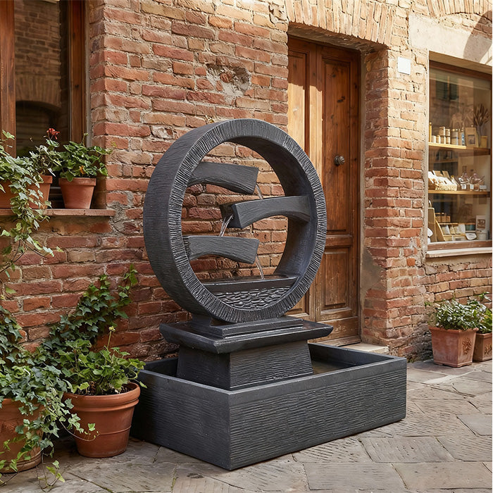 Round black outdoor water fountain with cascading tier design and rectangular basin, positioned against a rustic brick patio wall with potted plants.