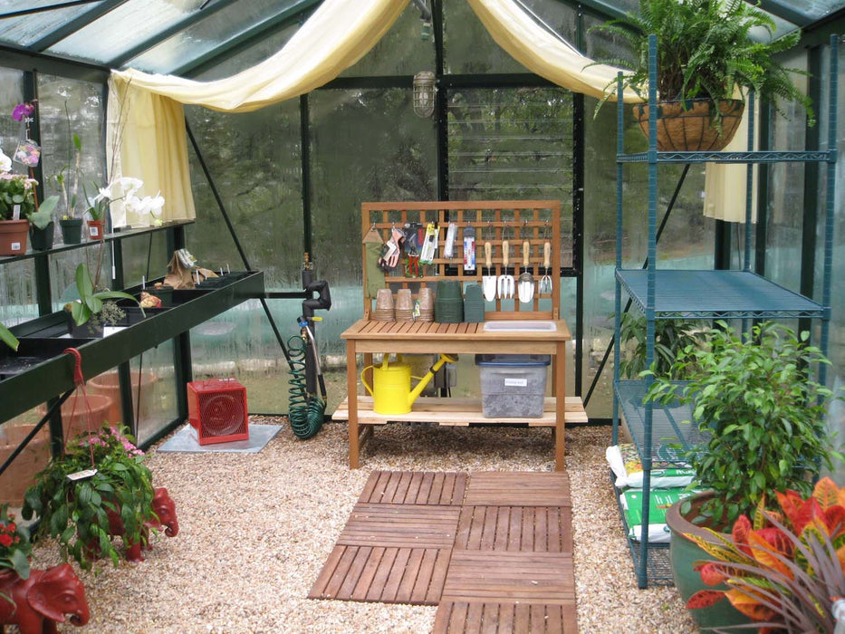 Inside the Royal Victorian VI 34 Greenhouse featuring a wooden potting bench, gardening tools, potted plants, and gravel flooring.