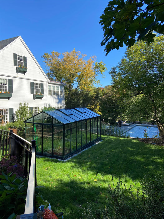 Side yard view of Royal Victorian VI 36 Greenhouse surrounded by grass, positioned near a home and swimming pool with trees in the background.