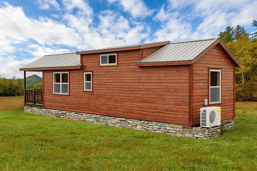 A brown, Pocono cabin with a gray metal roof sits on a grassy field. A small porch is visible on the left side, and there are several windows along the side of the cabin. Trees and mountains are visible in the background under a blue sky with white clouds.