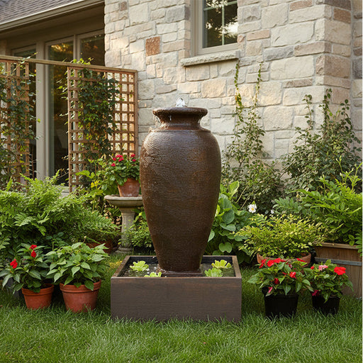 Rustic amphora outdoor water fountain with square basin, textured finish and floating plants in landscaped garden setting.