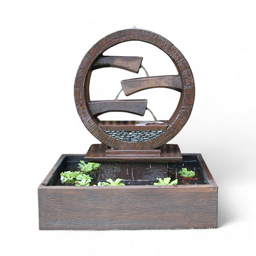 A rustic brown Wagon Wheel Fountain by Exaco, featuring water flowing over three tiers within a circular design, with small green aquatic plants floating in the rectangular basin
