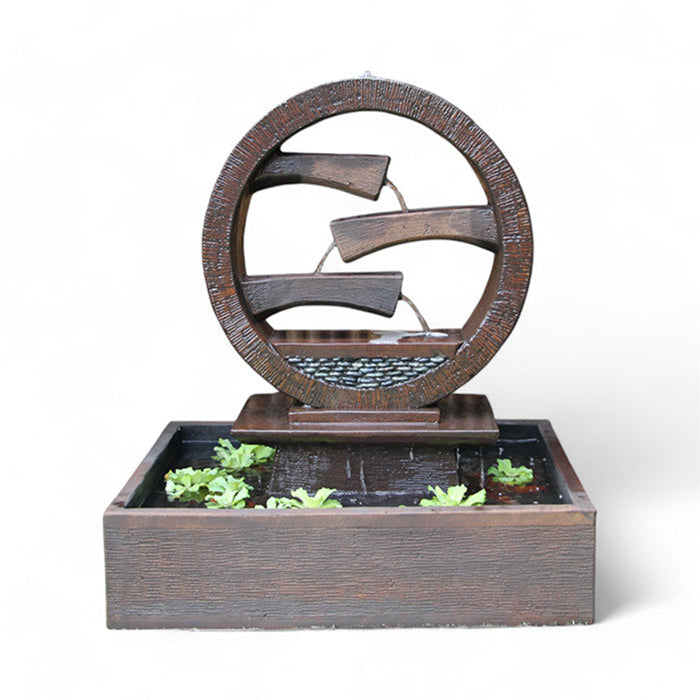 A rustic brown Wagon Wheel Fountain by Exaco, featuring water flowing over three tiers within a circular design, with small green aquatic plants floating in the rectangular basin