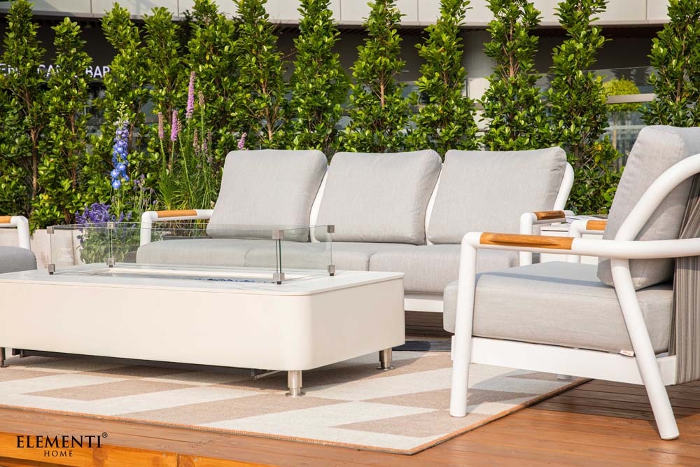 An Elementi Outdoor Furniture Santorini Collection seating group, including a sofa, two chairs, and a fire table, on a striped rug.