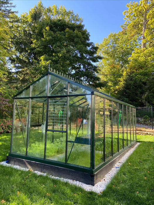 Side angle view of Exaco Janssens Royal Victorian VI 36 Greenhouse on a white stone base, showcasing clear glass walls and lush plants inside.