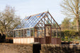 Side view of the cedar Westminster greenhouse structure, set in a scenic garden surrounded by trees and paved walkways.