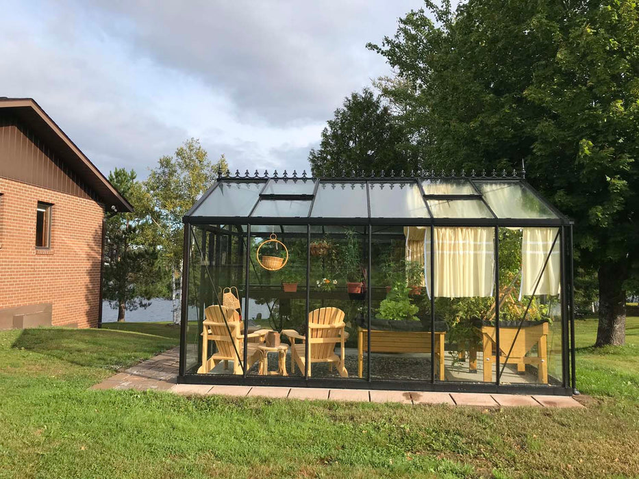 Side view of Royal Victorian VI 34 Greenhouse with curtain shades and wooden seating, set in a spacious yard beside a brick house.