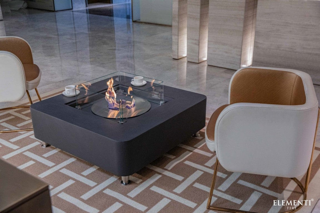 Elementi Perth Ethanol Fire Table in slate black shown in a modern room setting with white chairs and coffee cups