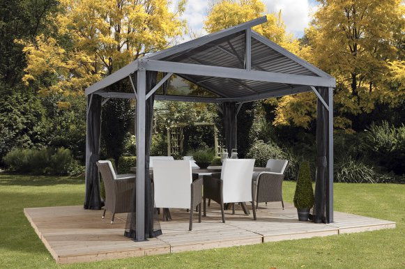 Sojag Sanibel II 10x10 gazebo with mosquito netting on a wooden deck. Outdoor furniture sits in the shade of the gazebo, with trees in the background