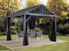 ojag South Beach I Hardtop Gazebo 12x12 ft in Light Gray, set up on a wooden deck in a lush garden, showcasing the spacious and elegant outdoor shelter with dining furniture.