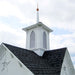 Close-up of Little Cottage Co. Star Barn cupola featuring white arched vents, copper spire tip, and dark shingled roof under blue sky.