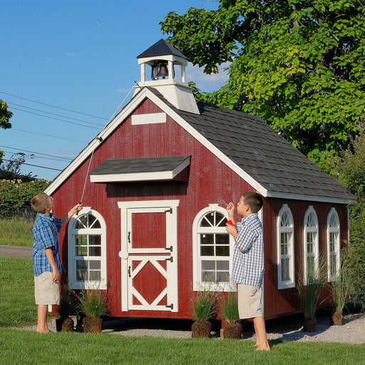 Two children playing outside the Stratford Schoolhouse playhouse from Little Cottage Co., featuring a red exterior, bell tower, and white trim windows