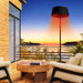 The standing heater is on a balcony against a backdrop of a stunning sunset over a coastal landscape, highlighting its ambient lighting feature.