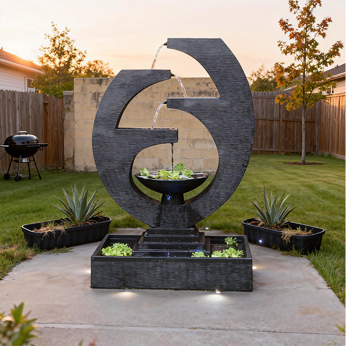 Eclipse outdoor water feature at sunset with illuminated base lights, circular design and cascading water in landscaped backyard patio.
